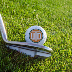 28th Annual UT Dallas Scholarship Golf Tournament. A golf ball bearing the UTD logo, about to be hit by a golf club.