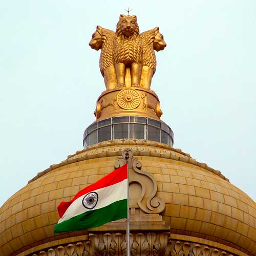 The Indian flag and the State Emblem atop Vidhana Soudha in Bangalore. Photo Credit: CC BY 3.0 2005 Mellisa Anthony Jones [https://commons.wikimedia.org/wiki/File:India_flag_emblem.jpg]