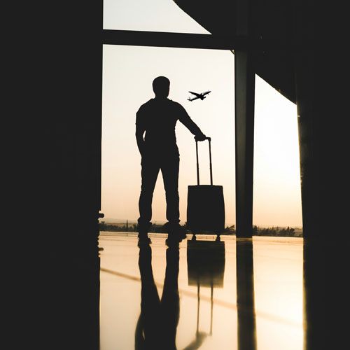 A traveler with a piece of rolling luggage looks out of the full-length window of an airport terminal at a plane rising into a dawn sky whose light casts everything into silhouette while reflecting it onto the polished floor. Photo Credit: Yousef Alfuhigi on Unsplash [https://unsplash.com/photos/silhouette-of-man-holding-luggage-inside-airport-bMIlyKZHKMY]