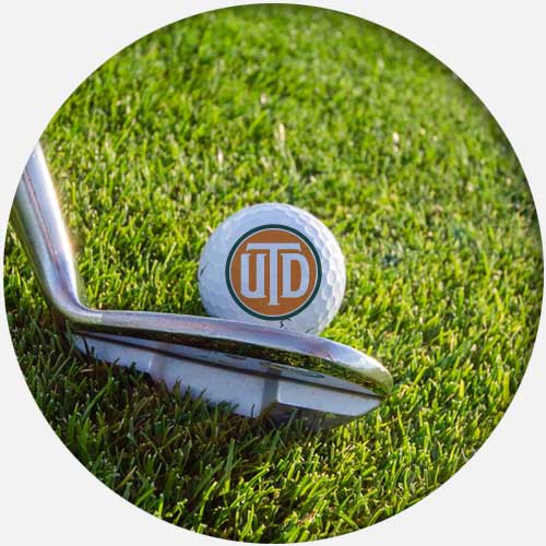 The end of a golf club pressed up against a golf ball bearing the UTD logo.